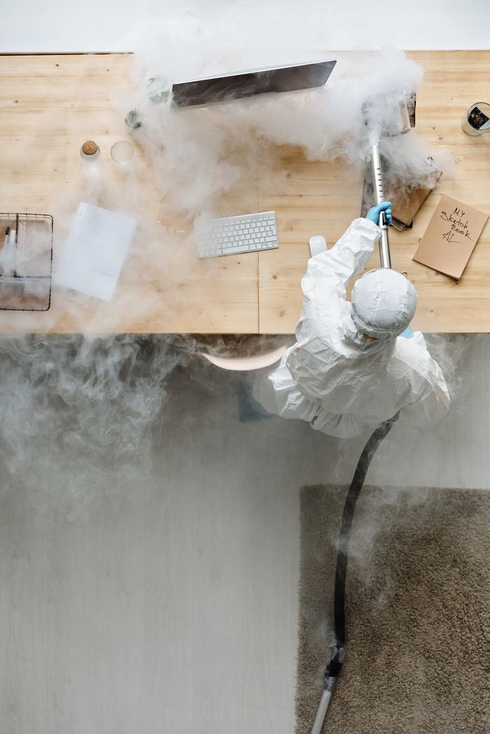 services-01 A person in protective gear sanitizing an office desk using fumigation equipment during daytime.