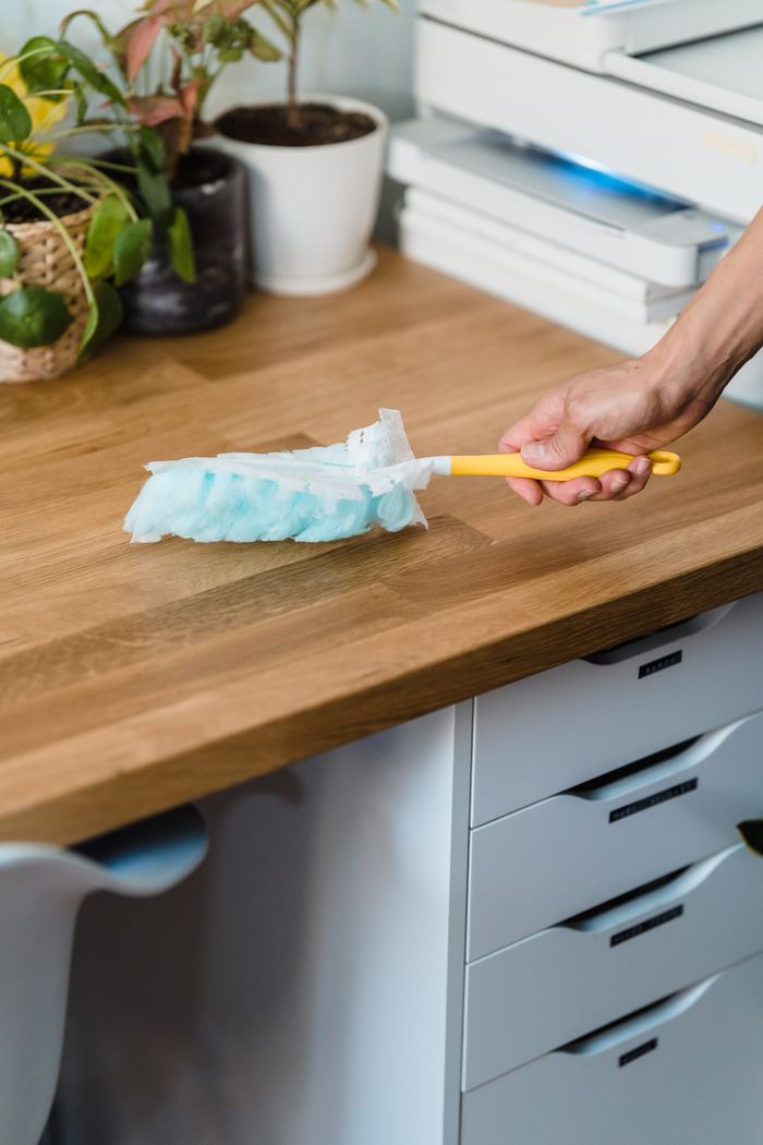 about-01 A hand using a feather duster to clean a wooden desk, surrounded by plants and office items.